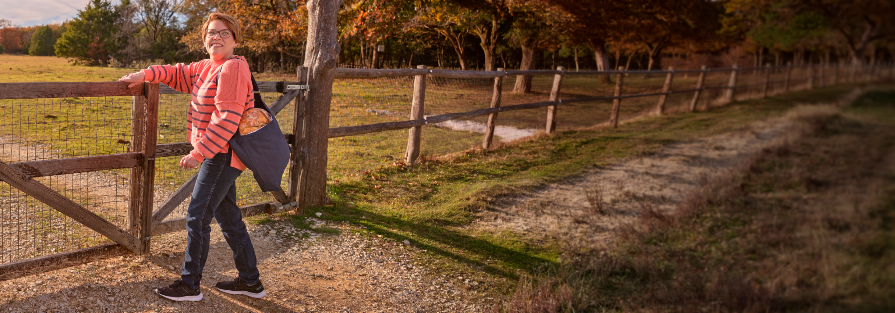 Karon opens a gate in a farm field with a loaf of bread in a bag slung over her shoulder.