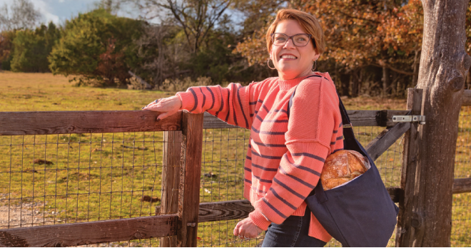 Karon opens a gate in a farm field with a loaf of bread in a bag slung over her shoulder.