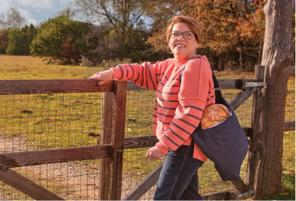 Karon opens a gate in a farm field with a loaf of bread in a bag slung over her shoulder.