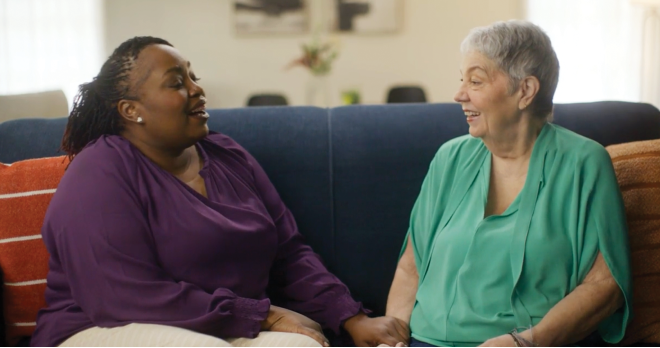 Ebony and Pam sit on a blue couch chatting.