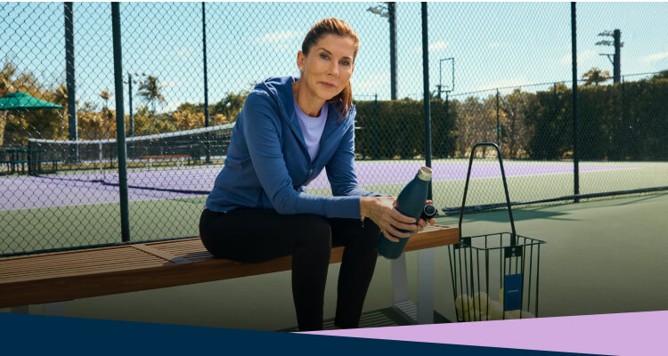 Tennis legend Monica Seles sits on a bench next to a tennis court.