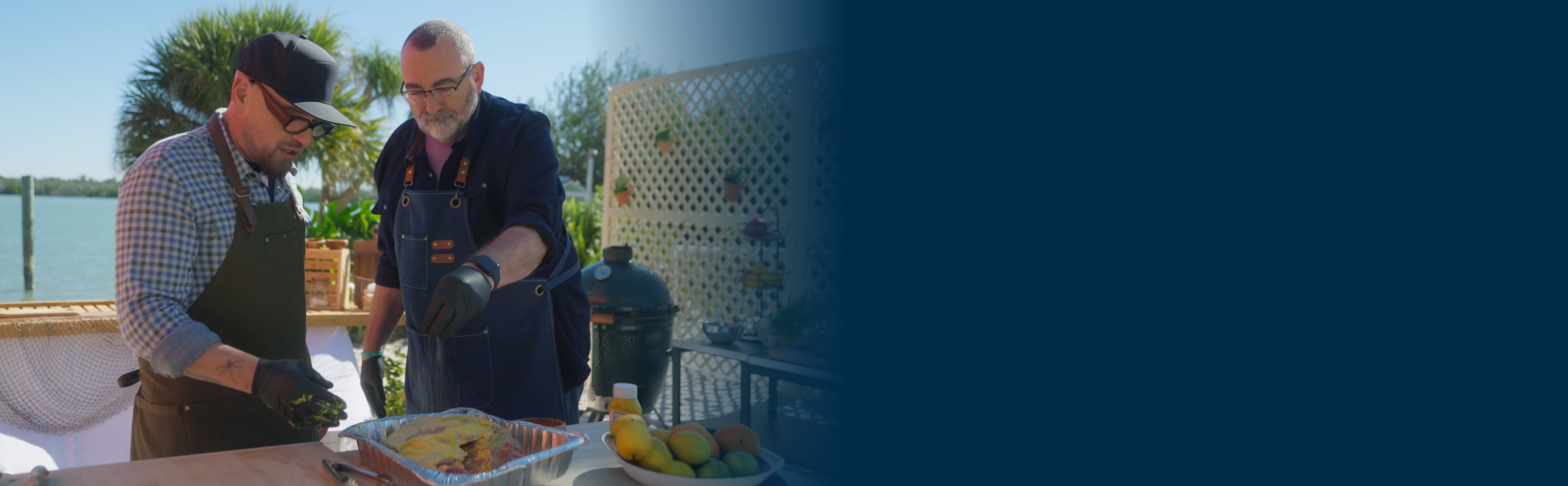 Steve and chef Michael Symon prepare pork shoulder in an outdoor kitchen alongside the ocean.
