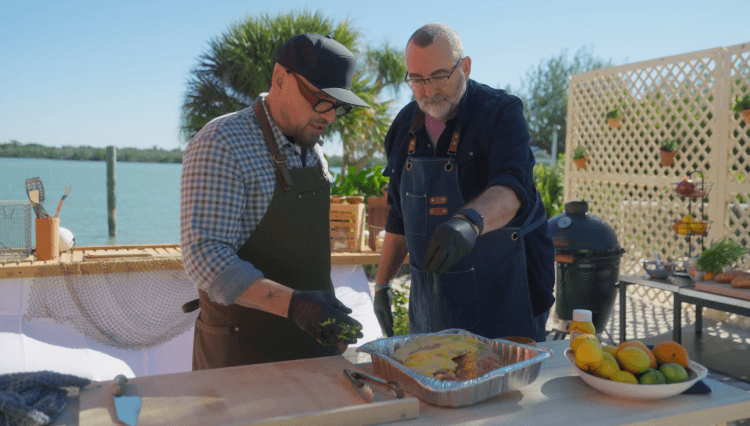 Steve and chef Michael Symon prepare pork shoulder in an outdoor kitchen alongside the ocean.
