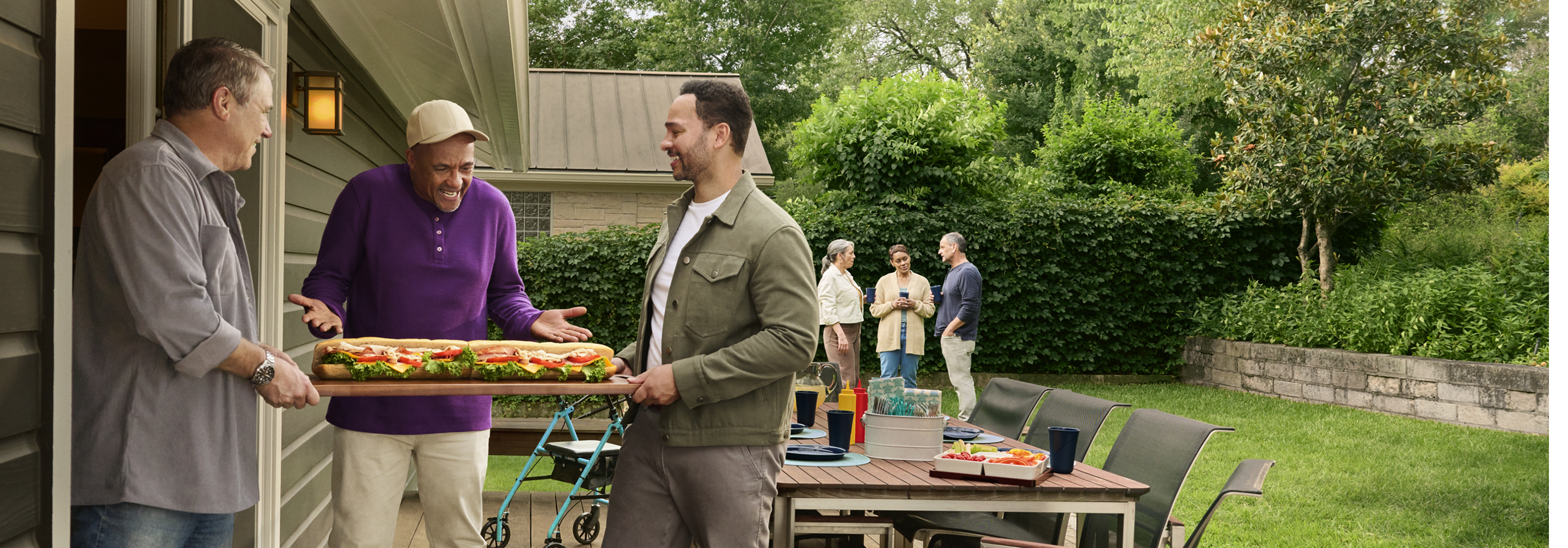 A group of men bringing a large sandwich to a backyard cookout
